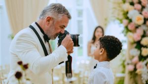 An experienced wedding photographer teaching a young mentee about camera settings during a live wedding shoot, with a picturesque ceremony backdrop.