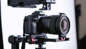 Close-up of a Canon RF lens on a mirrorless camera video rig with follow-focus and shotgun microphone, lit by soft studio light, with a blurred softbox, tripod, and gimbal in the background.