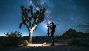 Photographer with tripod beside a weathered Joshua tree under a bright Milky Way in a moonless desert basin, faint red headlamp glow, distant mesas and dunes fading into the background.