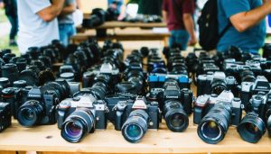"Various vintage and modern cameras and lenses on a wooden table at a photography swap meet with blurred background of people interacting."