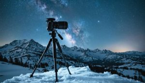 DSLR on a sturdy tripod in a snowy alpine landscape at night, lit by cool moonlight with sharp stars and a faint Milky Way, with distant pine trees and mountain silhouettes in the background.