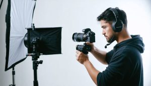 Videographer in a home studio adjusting a gimbal-mounted mirrorless camera with a shotgun mic under an LED softbox, wearing headphones, with a blurred tripod and editing workstation in the background