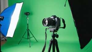 Cinema camera on tripod aimed at a seamless green screen with tracking markers, chrome and gray reference spheres on a stand, no actors, with softly blurred studio lights and flags in the background.