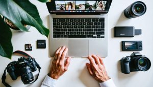 Wedding photographer at a minimalist desk using a laptop with camera, memory cards, and external SSD; a softly blurred second monitor shows a grid of wedding photo thumbnails; natural window light creates a calm, efficient atmosphere.