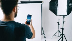 Photographer using a smartphone to control multiple smart LED panels and softboxes in a modern studio, with subtle blue and amber accent lighting and a seamless backdrop in the background.